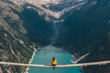 Woman sitting in the middle of a suspension bridge looking down at a mountain lake.