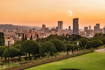 City skyline at sunset with high-rise buildings, a crescent moon overhead, and a green park with trees in the foreground.