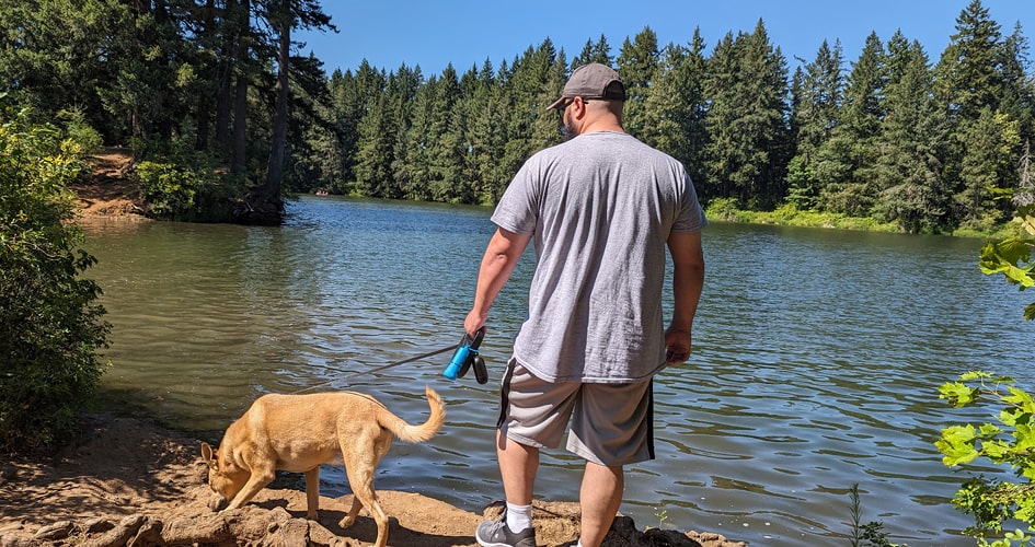 A photo of James Ortiz and Scooter, a Carolina dog crossbreed, outdoors near a body of water. Their backs are to the camera.