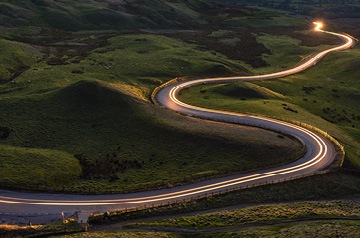 Headlights on a highway that snakes through green hills.