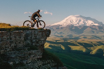 cyclist resting at cliff's edge, mountains behind