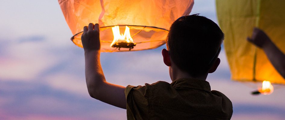 A child, seen from the back, prepares to set a floating lantern aloft.
