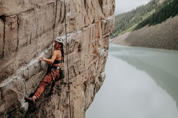 Woman in helmet using ropes to climb a canyon wall above a river.