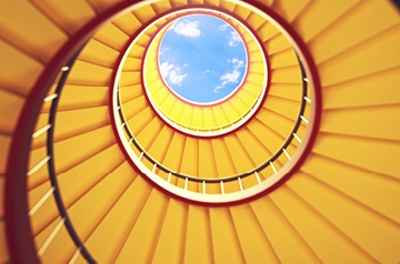 Upward view of a yellow spiral staircase circling a skylight with blue sky and clouds.